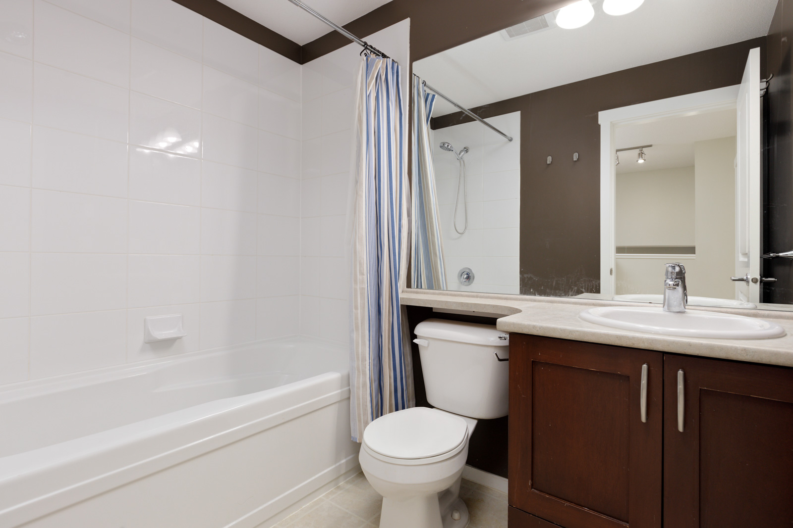 Modern bathroom with a bathtub, striped shower curtain, toilet, and a sink with a mirror above a wooden cabinet.