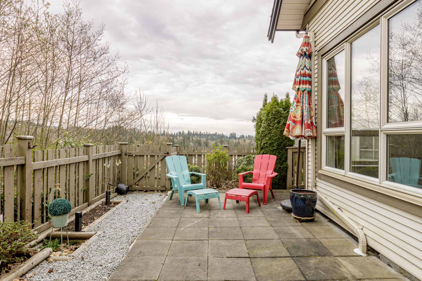 A patio with a fenced yard features three Adirondack chairs in blue and red, a large patio umbrella, and potted plants. Beyond the fence, there is a view of trees and hills under a cloudy sky.
