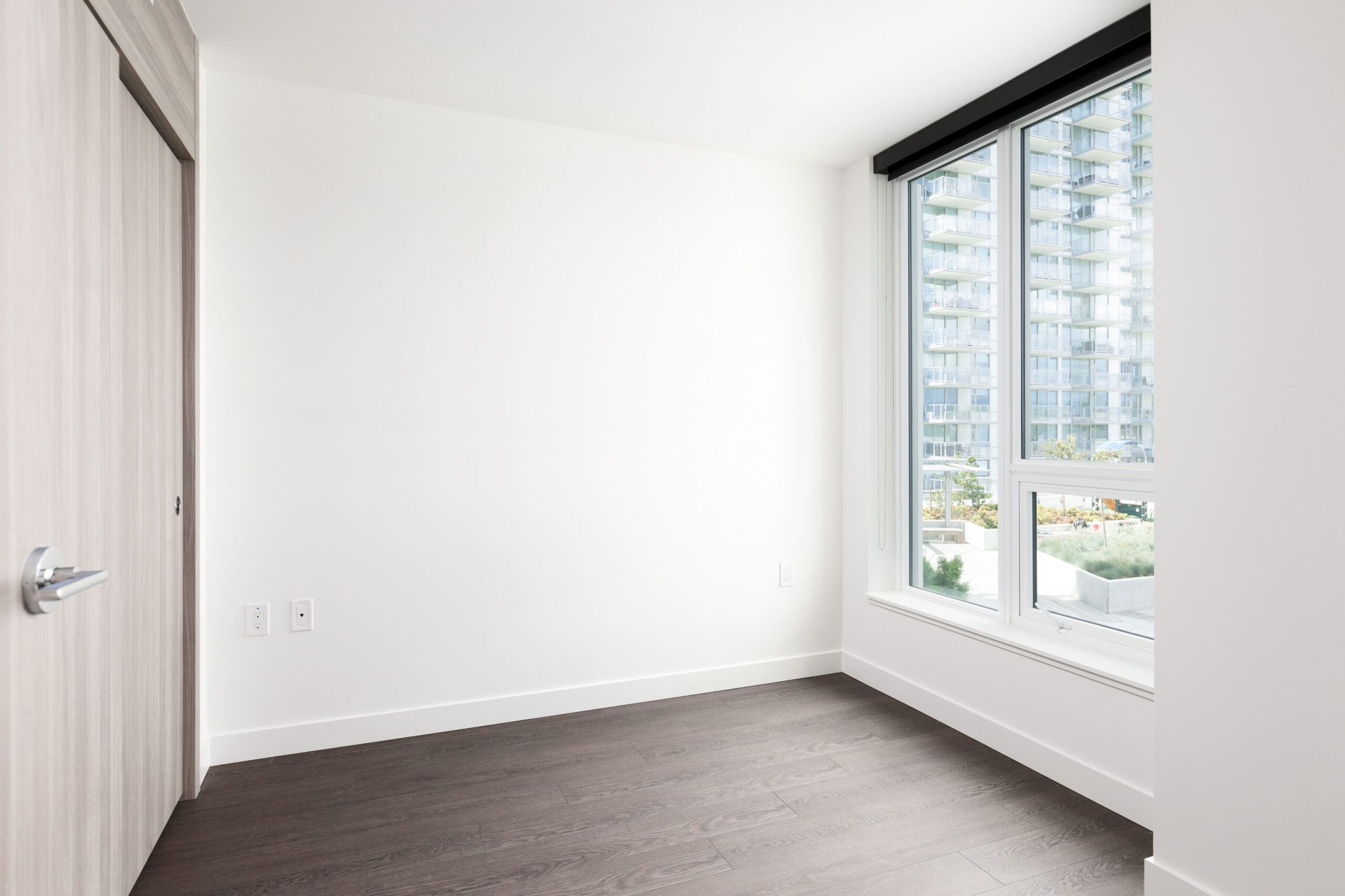 Empty room with white walls, dark wood flooring, a closed door, and a large window showing an exterior view of modern apartment buildings.