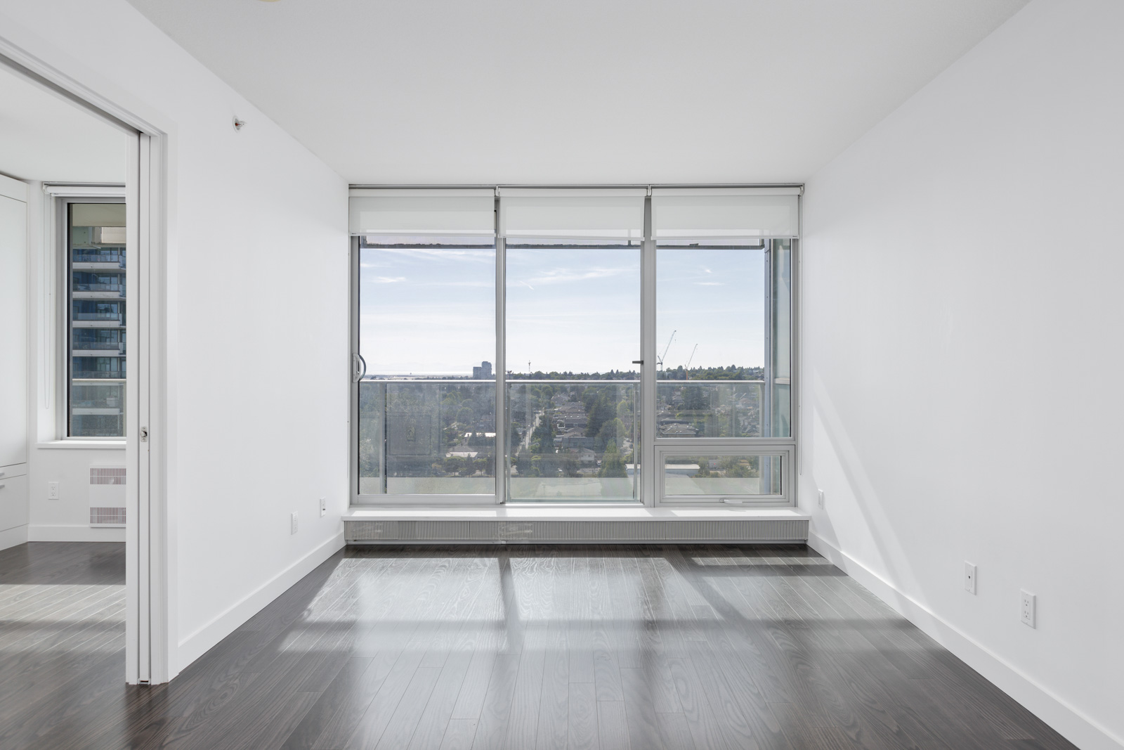 Bright, empty room with dark hardwood floors, large windows, and a view of the cityscape outside.
