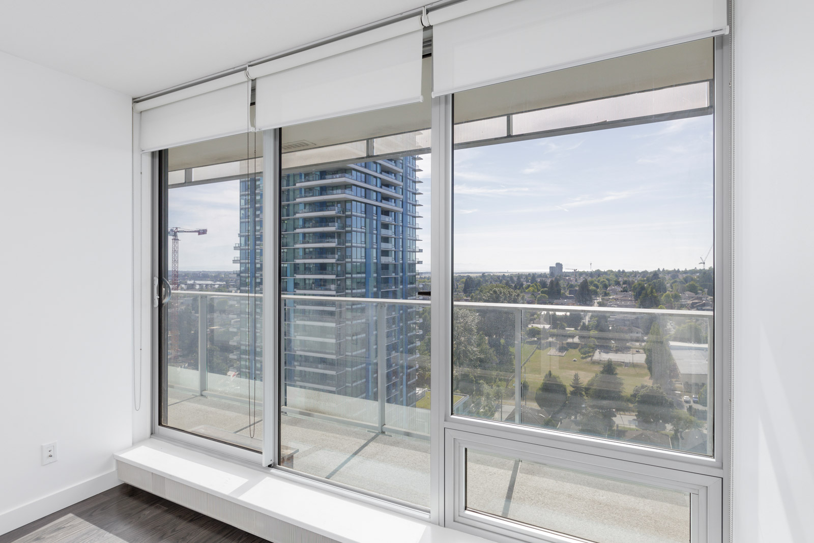 Modern apartment balcony with glass railing, overlooking a cityscape with a tall building and greenery in the background. Large sliding glass doors enhance the view from the interior.