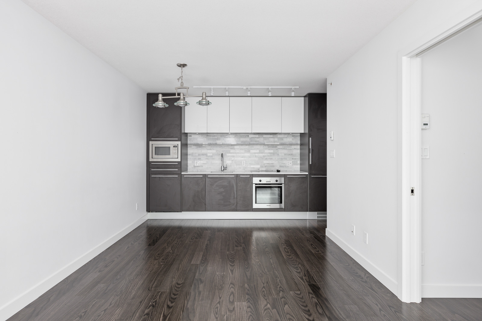 Modern, empty kitchen interior with white walls and dark hardwood floors. Features dark cabinets, a tiled backsplash, an oven, and a microwave. Ceiling lights hang above the space.