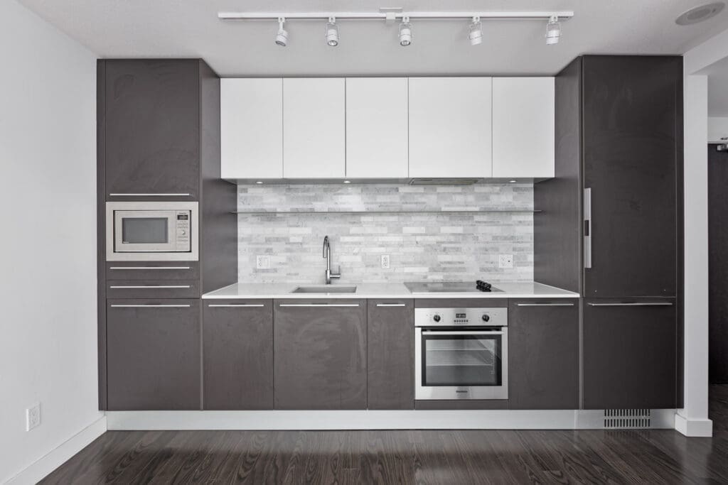 Modern kitchen with dark cabinets, white upper cupboards, stainless steel appliances, and a gray backsplash. Built-in oven and microwave are visible.
