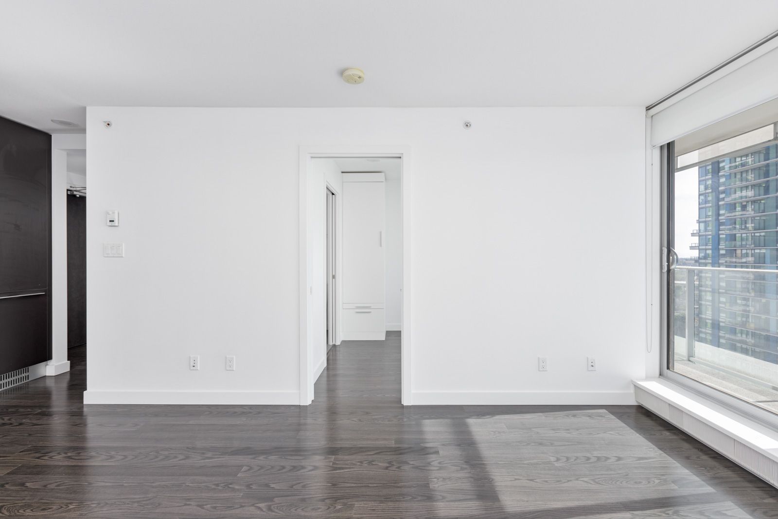 Empty modern room with white walls and dark wood flooring, large window with a view of city buildings on the right, and an open doorway leading to another room.
