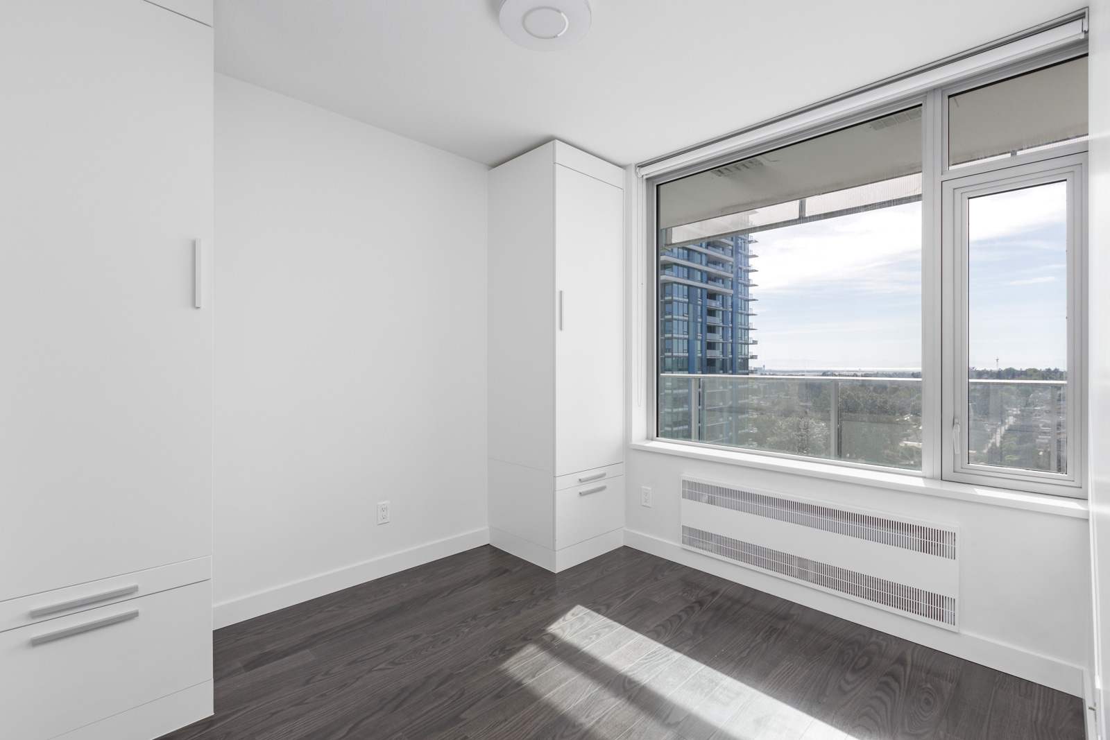 A bright, empty room with white walls, dark hardwood floors, a tall wardrobe, a large window overlooking a cityscape, and a wall-mounted radiator.
