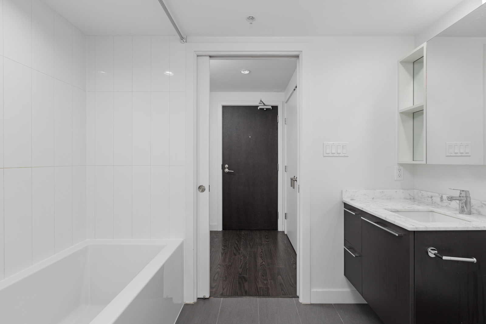 Modern bathroom with a white bathtub, dark wood vanity, and a marble countertop. Doorway opens to a hallway with dark wood flooring. White walls and minimal decor create a clean look.