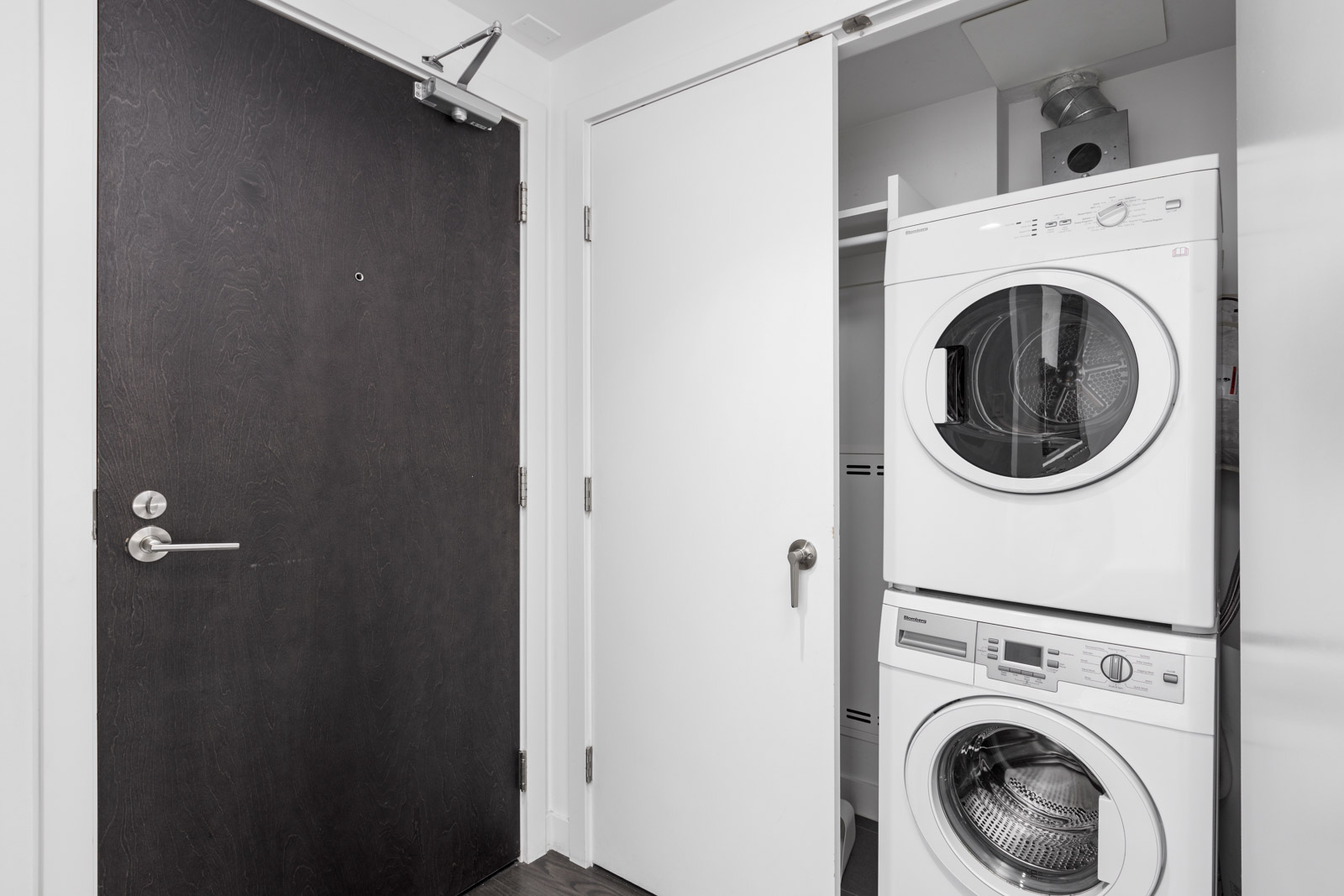 Stacked washer and dryer in a closet with sliding white doors, adjacent to a black entrance door in a modern laundry area.