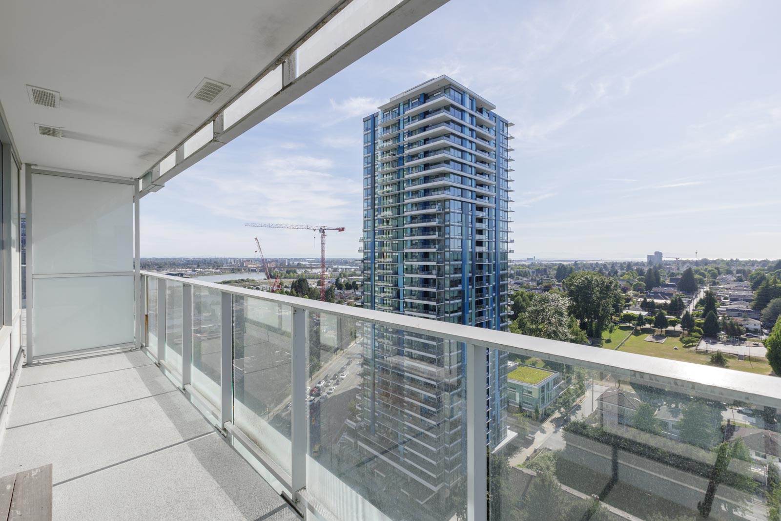 View from a balcony with glass railings showing a tall modern apartment building, construction cranes, and a cityscape under a clear sky.