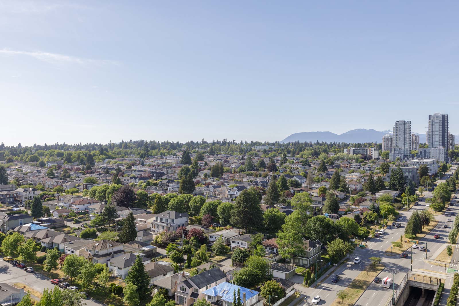 Aerial view of a residential neighborhood with green trees, houses, and roads. High-rise buildings and mountains are visible in the background under a clear blue sky.