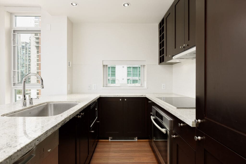 Modern kitchen with dark wood cabinets, white granite countertops, stainless steel appliances, and a sink. A small window provides natural light.