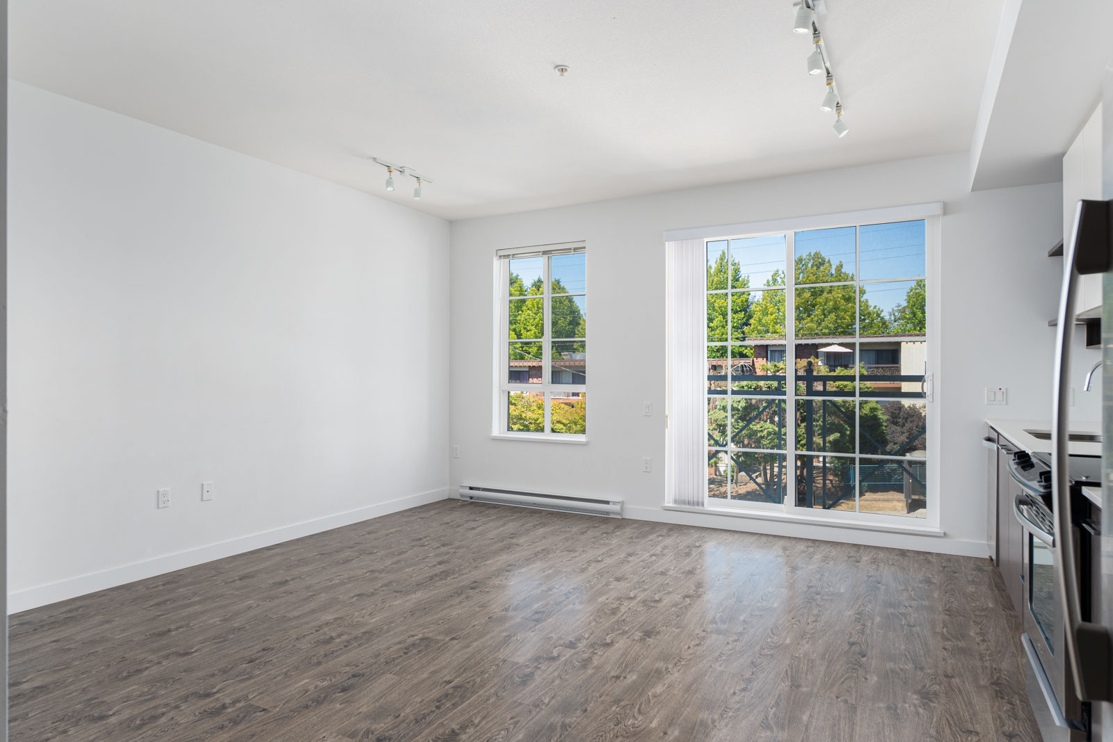 Empty room with white walls, wooden floor, large window, and track lighting. Kitchen area with a stainless steel stove on the right. Bright natural light illuminates the space.