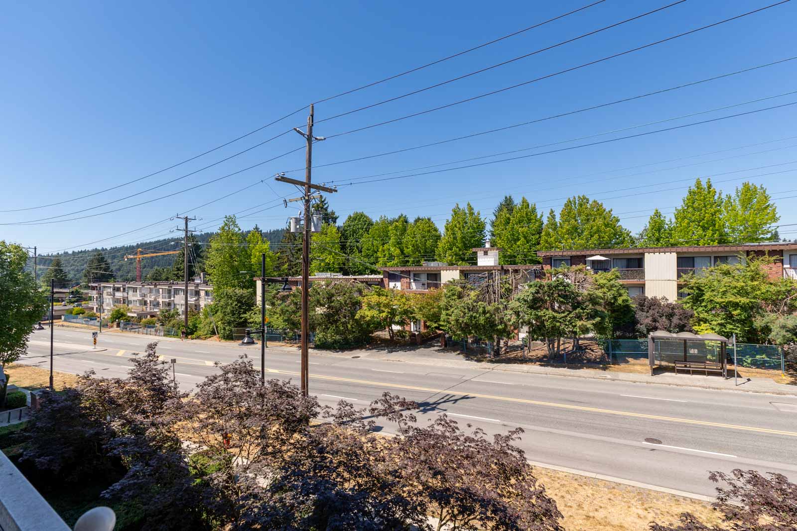 Street view with power lines, a two-lane road, a bus stop, and apartment buildings surrounded by greenery under a clear blue sky.