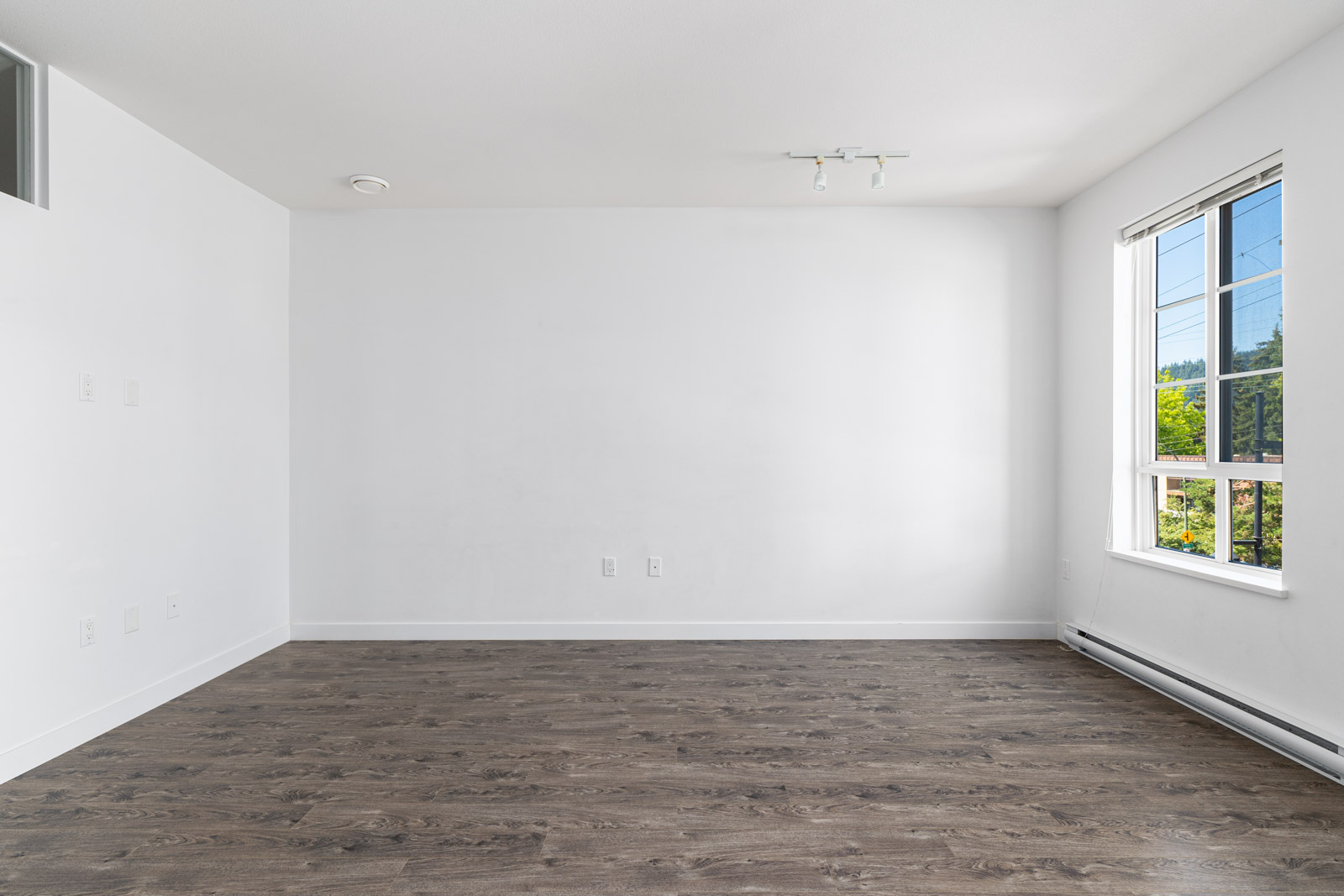 Empty room with white walls, wooden floor, and a large window on the right letting in natural light.