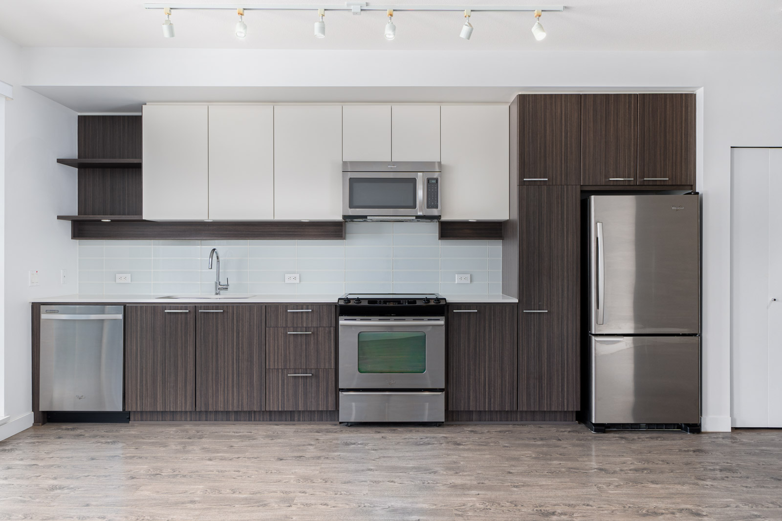 Modern kitchen with wood cabinets, stainless steel appliances, including a refrigerator, oven, microwave, and dishwasher, light blue backsplash, and track lighting above.