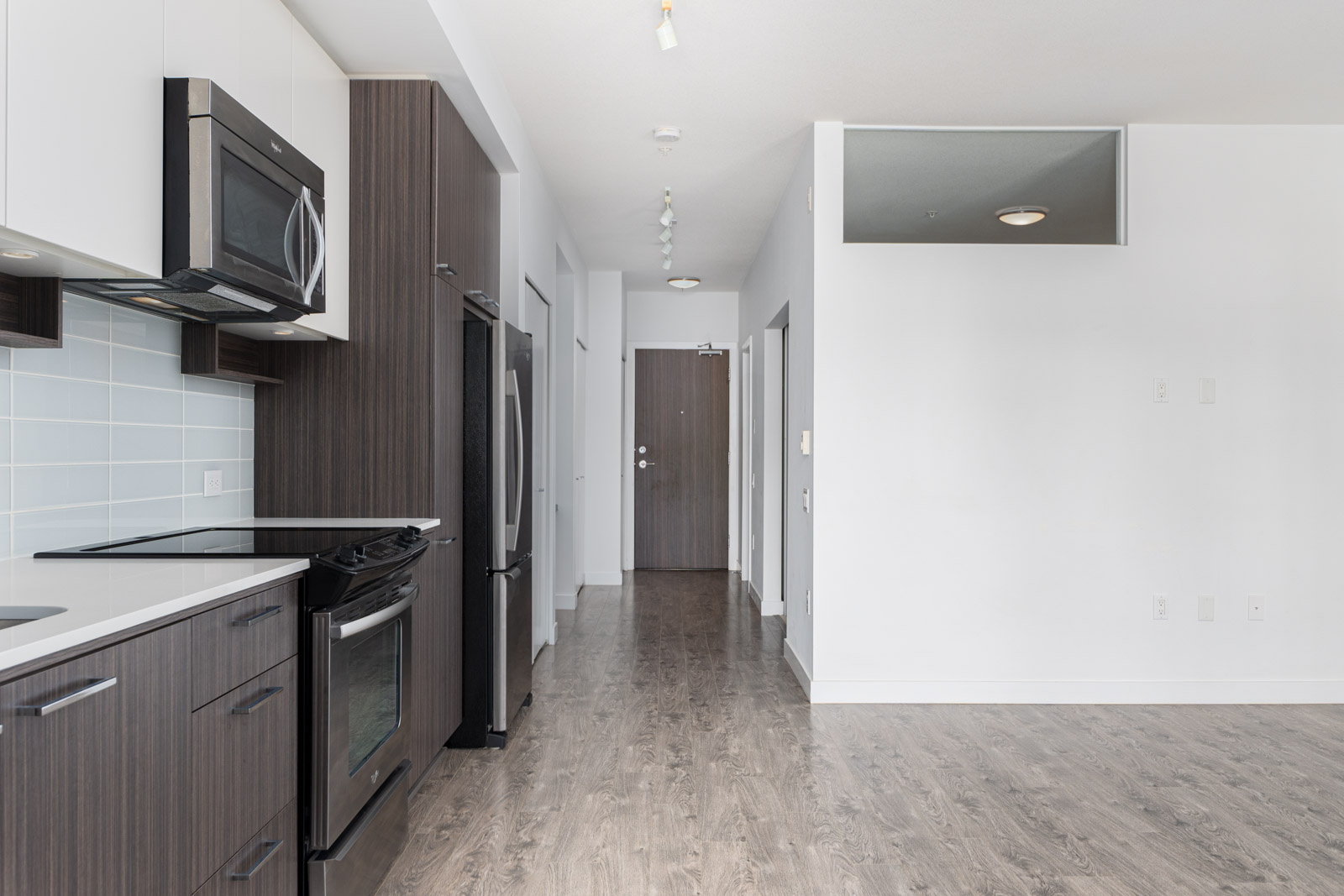 Modern kitchen and living area with dark wood cabinetry, stainless steel appliances, and light wood flooring. A hallway leads to a closed door.