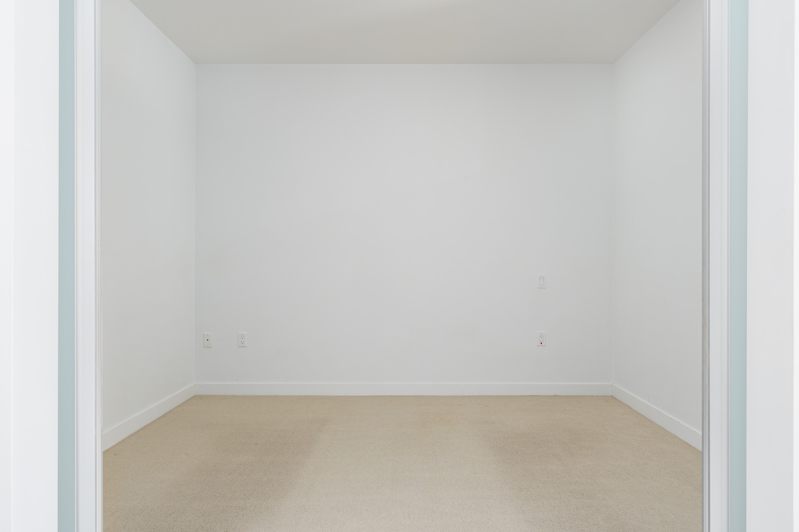 Empty white room with beige carpet, white walls, and a white ceiling. There are electrical outlets on the walls.