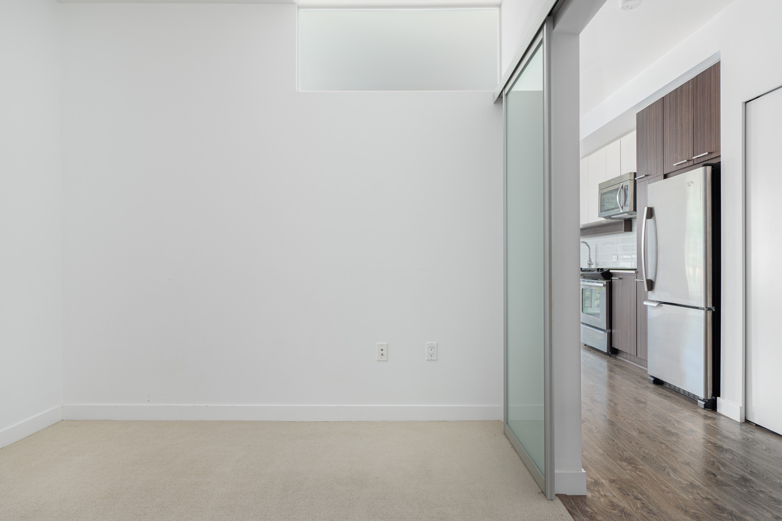 A partially visible modern kitchen with stainless steel appliances and hardwood floors, adjacent to a carpeted area with a white wall and a sliding glass door.