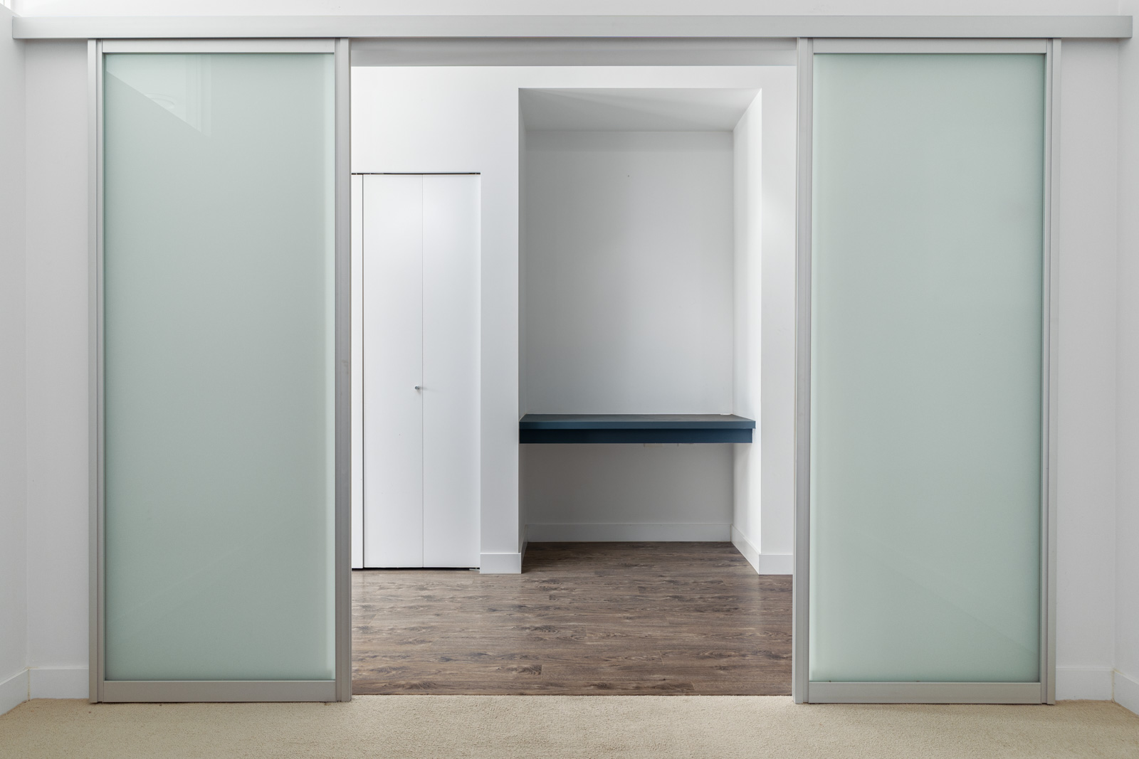 A room with light-colored walls and frosted glass sliding doors, featuring a small blue shelf mounted on the back wall, and a closet door on the left.