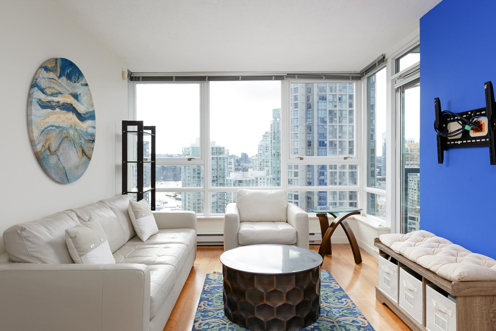 A modern living room with white furniture, a round coffee table, and a large blue accent wall. Large windows offer a cityscape view. A circular abstract painting hangs on the wall.