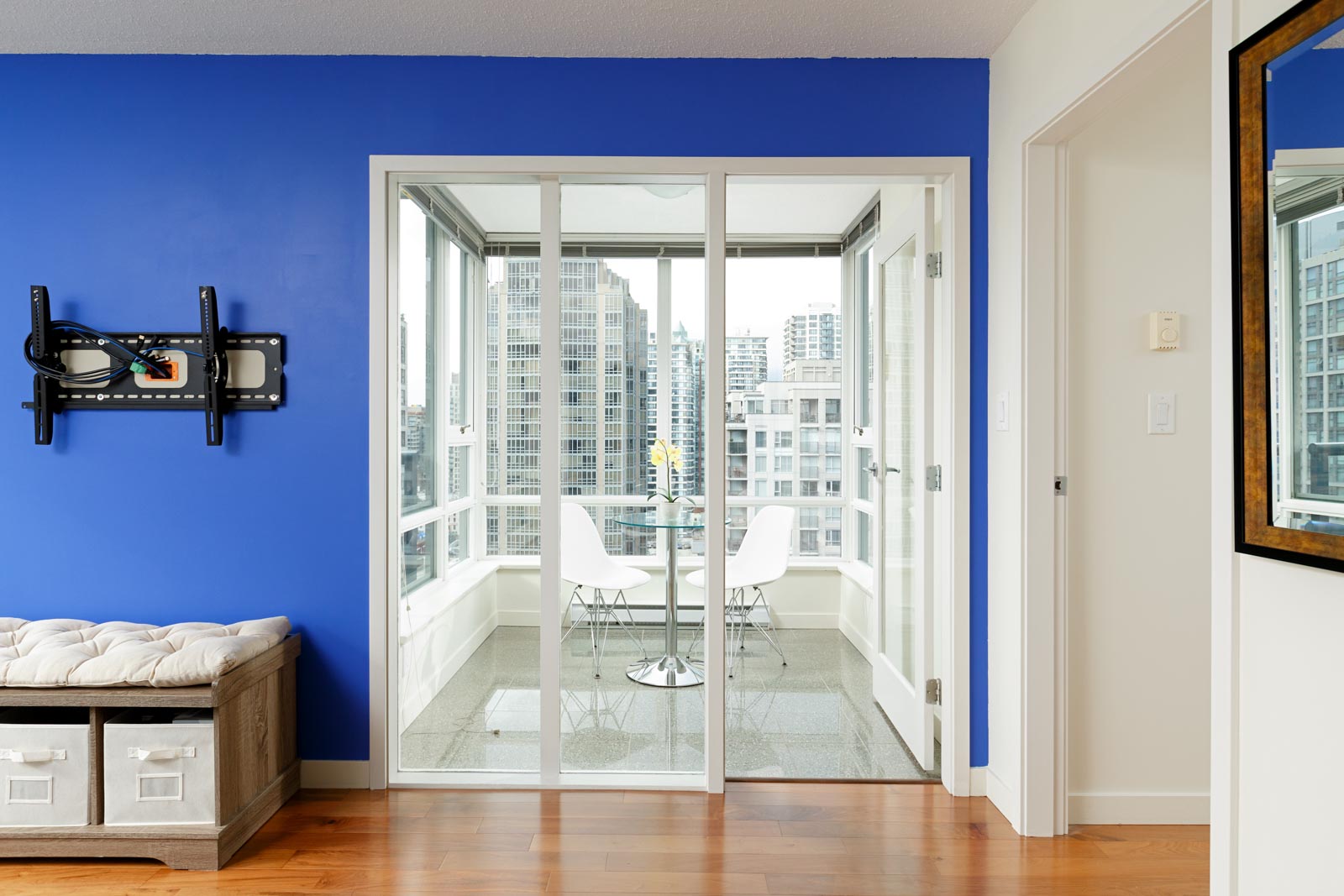 A modern apartment with blue accent wall features a seating area with a storage bench and wall-mounted TV bracket, opening to a balcony with a glass dining table and chairs overlooking city buildings.