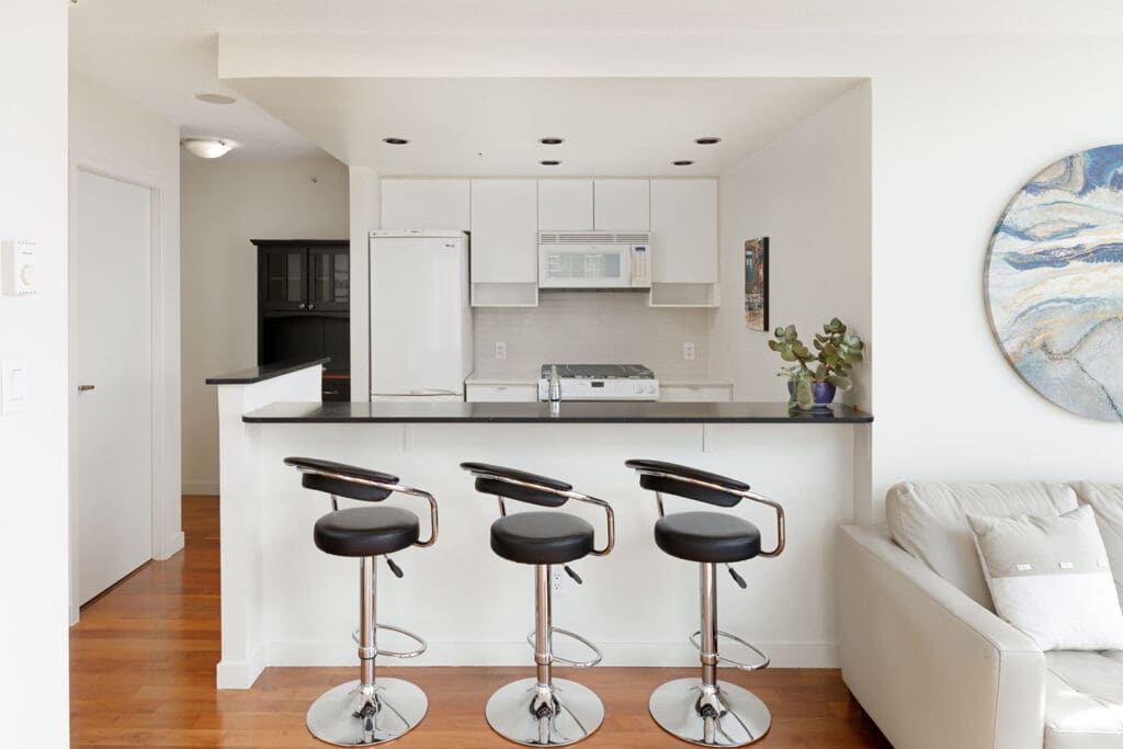 A modern kitchen with white cabinets, a white refrigerator, a microwave, and three black barstools along a black countertop. A gray couch and a wall painting are visible on the right.