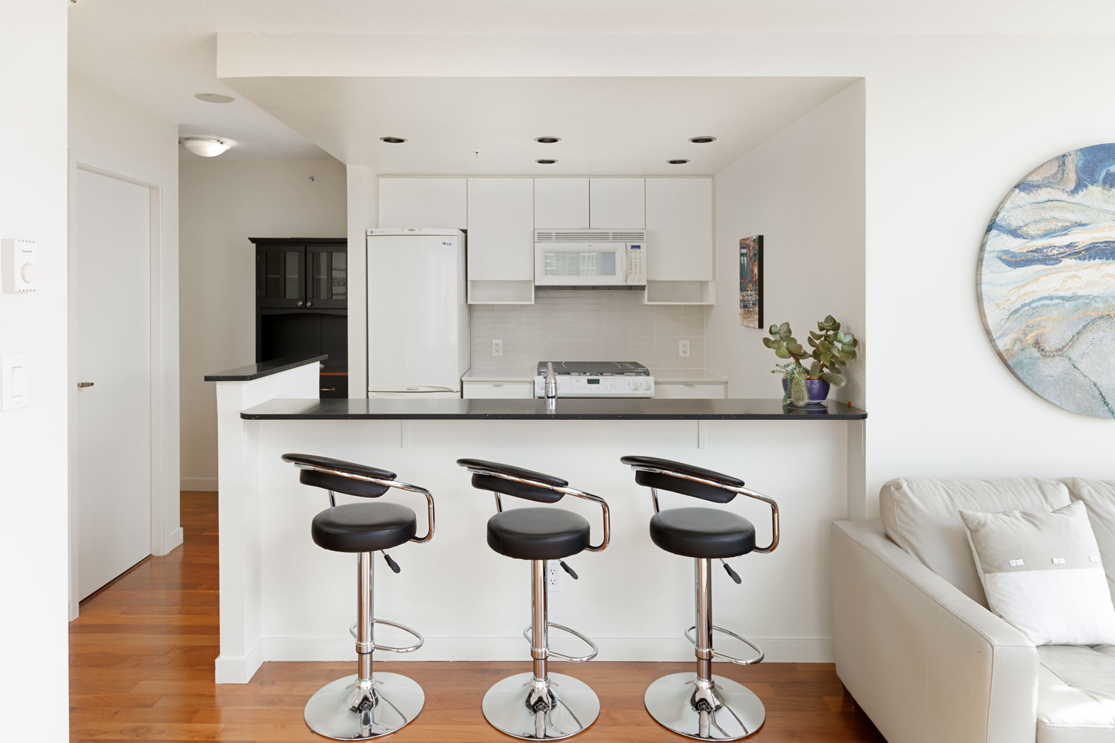 A modern kitchen with white cabinets, a white refrigerator, a microwave, and three black barstools along a black countertop. A gray couch and a wall painting are visible on the right.