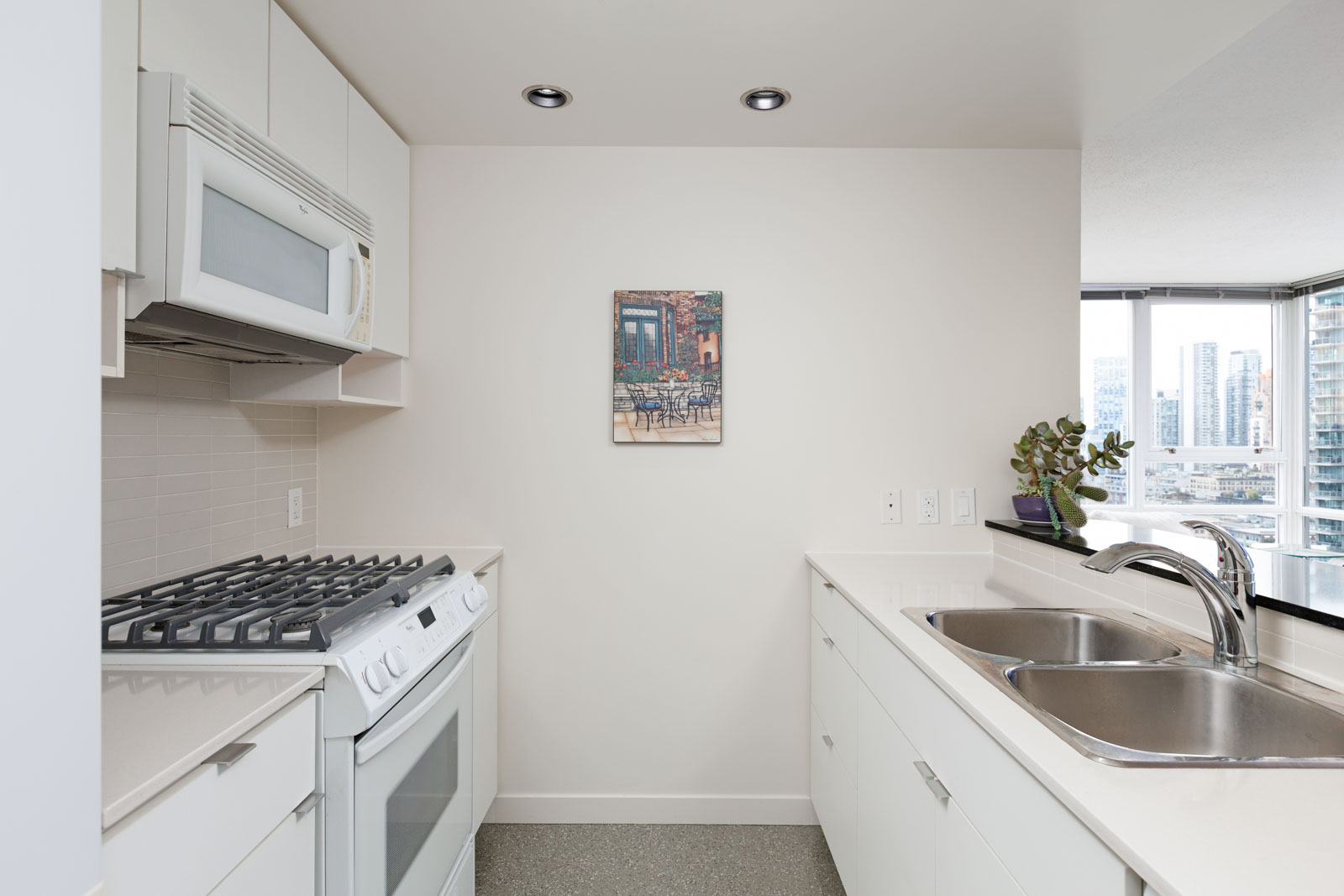 Modern kitchen with white cabinetry, gas stove, microwave, dual sink, and large windows. A framed picture of an outdoor patio hangs on the wall.
