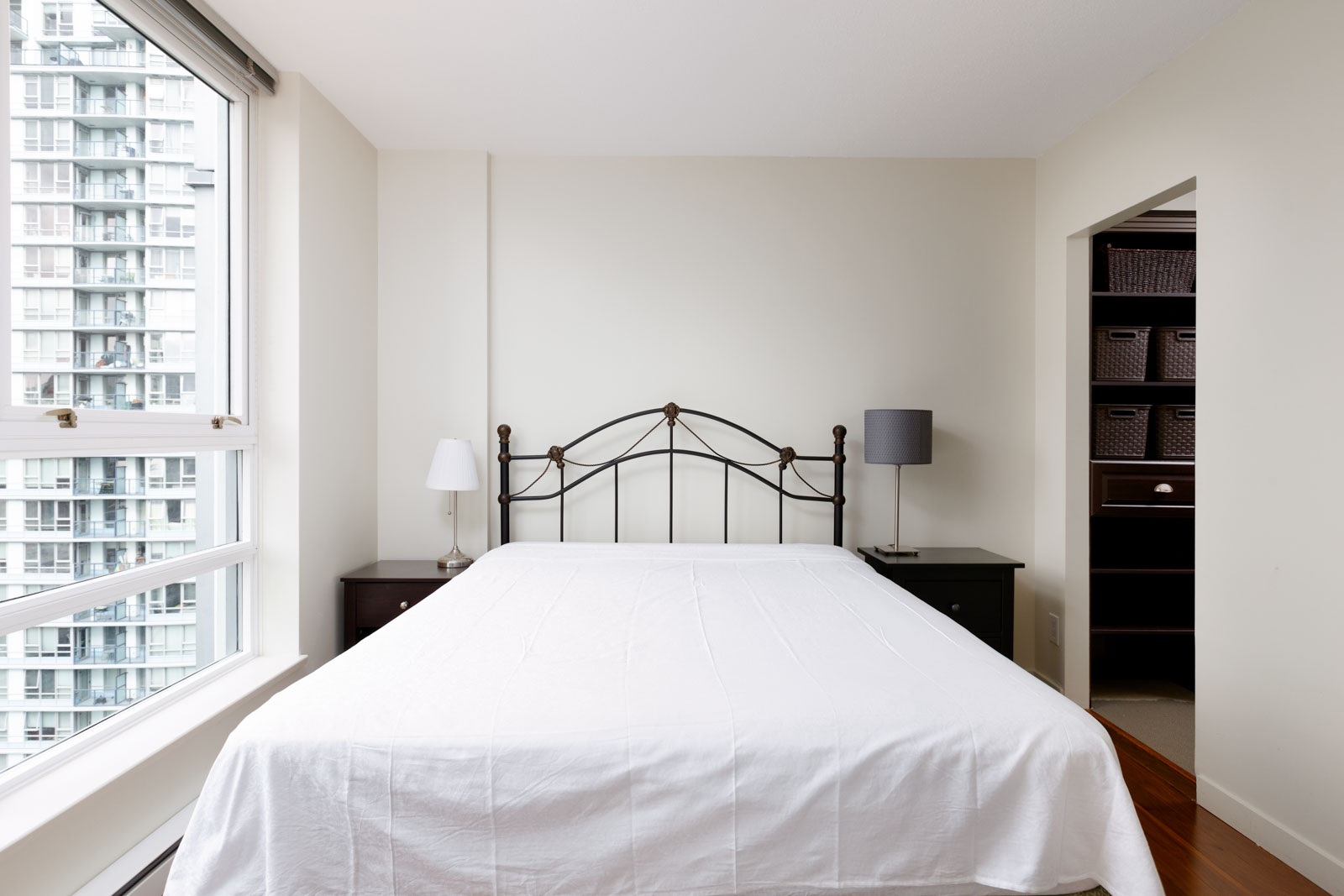 A bedroom with a white bed, metal headboard, two bedside tables with lamps, a window on the left, and an open closet with shelves on the right.