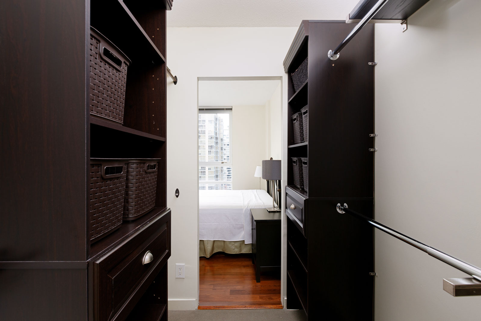 View of a walk-in closet with dark wood shelving and woven baskets, leading to a bedroom with a bed, nightstand, and a lamp, and a window showing an urban cityscape.