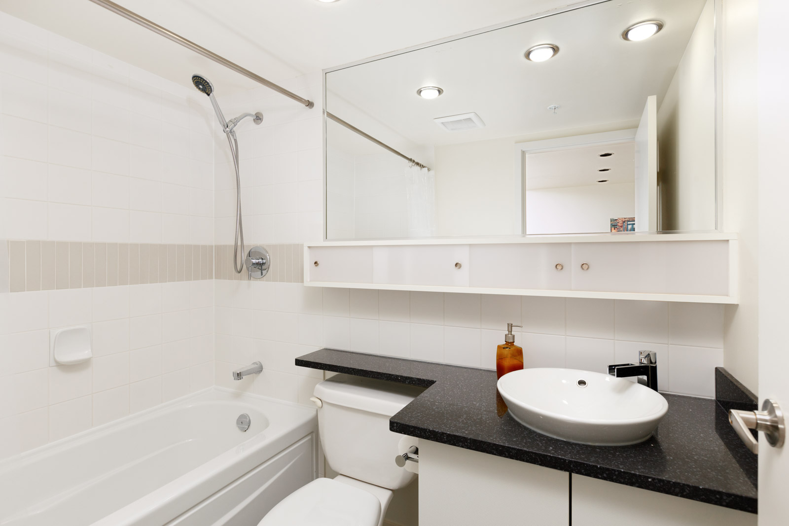 A modern bathroom with a bathtub, shower, white countertop, vessel sink, toilet, large mirror, and under-sink storage. The decor is minimalistic with white tiles and black countertop.