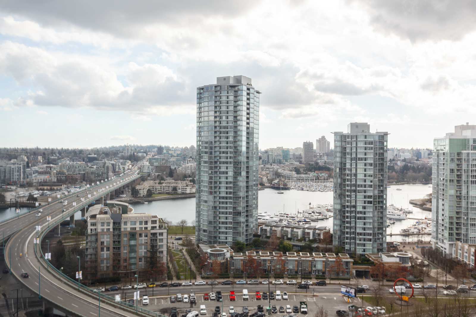 A cityscape view featuring high-rise buildings, a bridge on the left connecting to the buildings, and a marina with boats in the background under a cloudy sky.