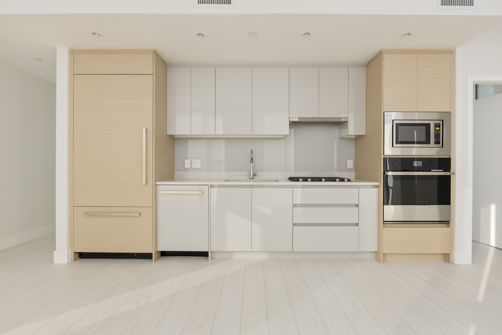 A modern kitchen with light wood cabinetry, white countertops, built-in refrigerator, oven, microwave, dishwasher, and stovetop. Walls are white, and the floor has light wood finish.