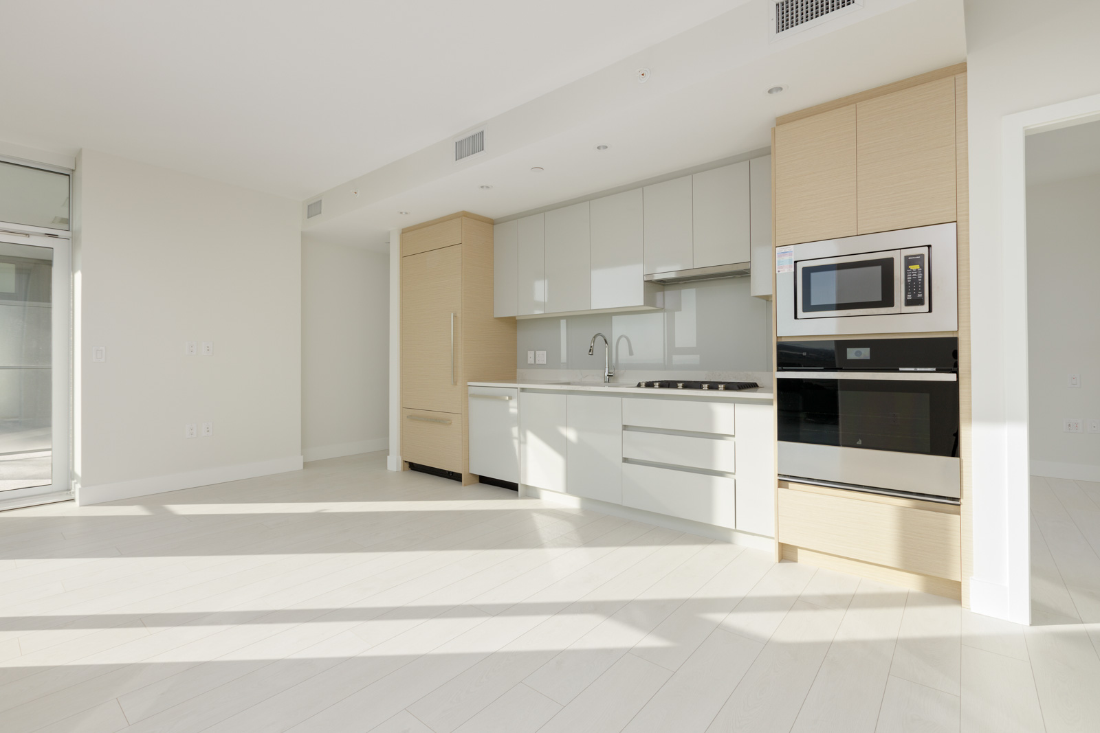 Modern kitchen with light wood and white cabinetry, built-in appliances, a sink, and a large window providing ample natural light. The space has minimalistic design and light-colored flooring.