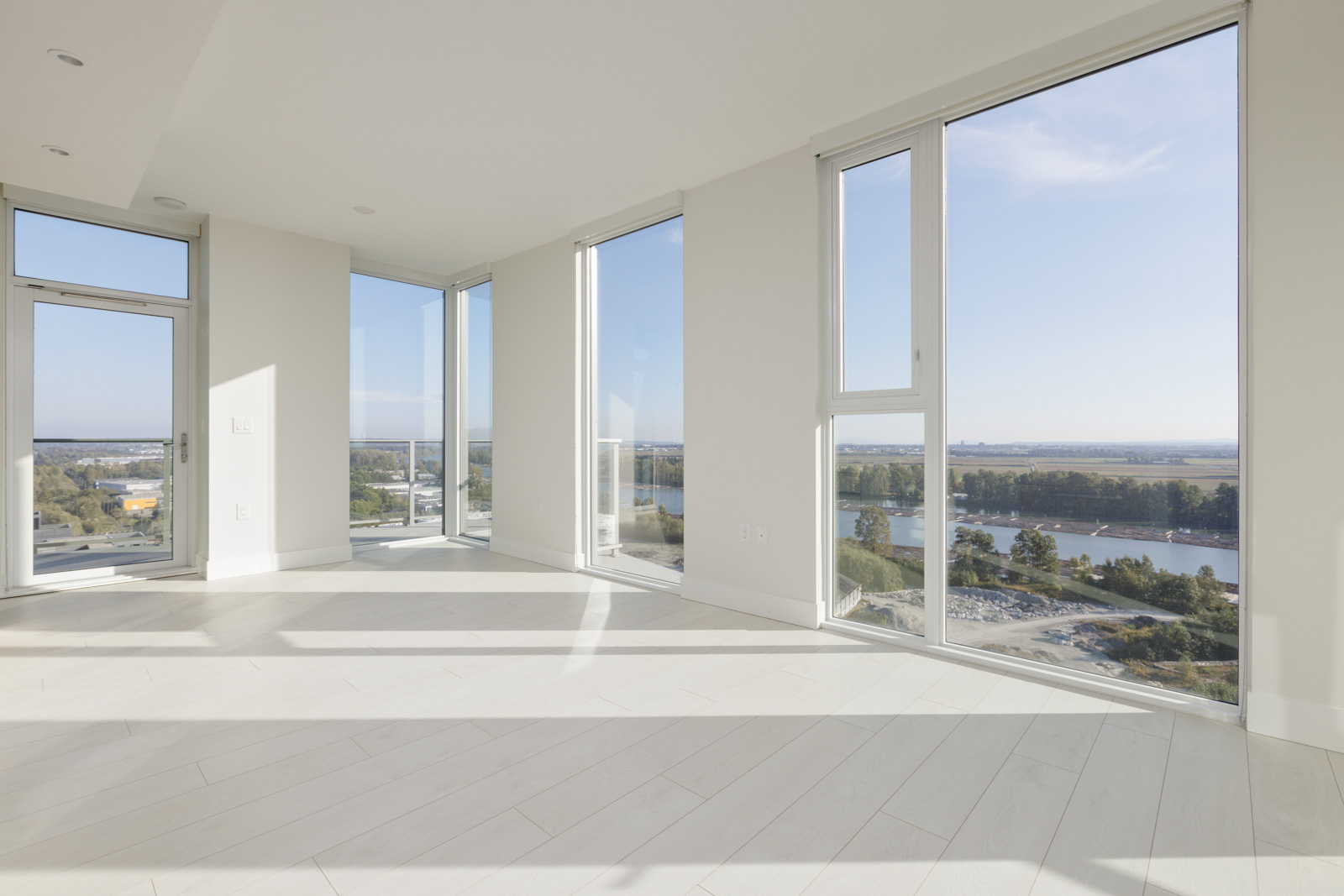 A spacious empty room with large floor-to-ceiling windows overlooking a river, surrounding landscape, and a partially visible balcony door. The room has light wood flooring and white walls.