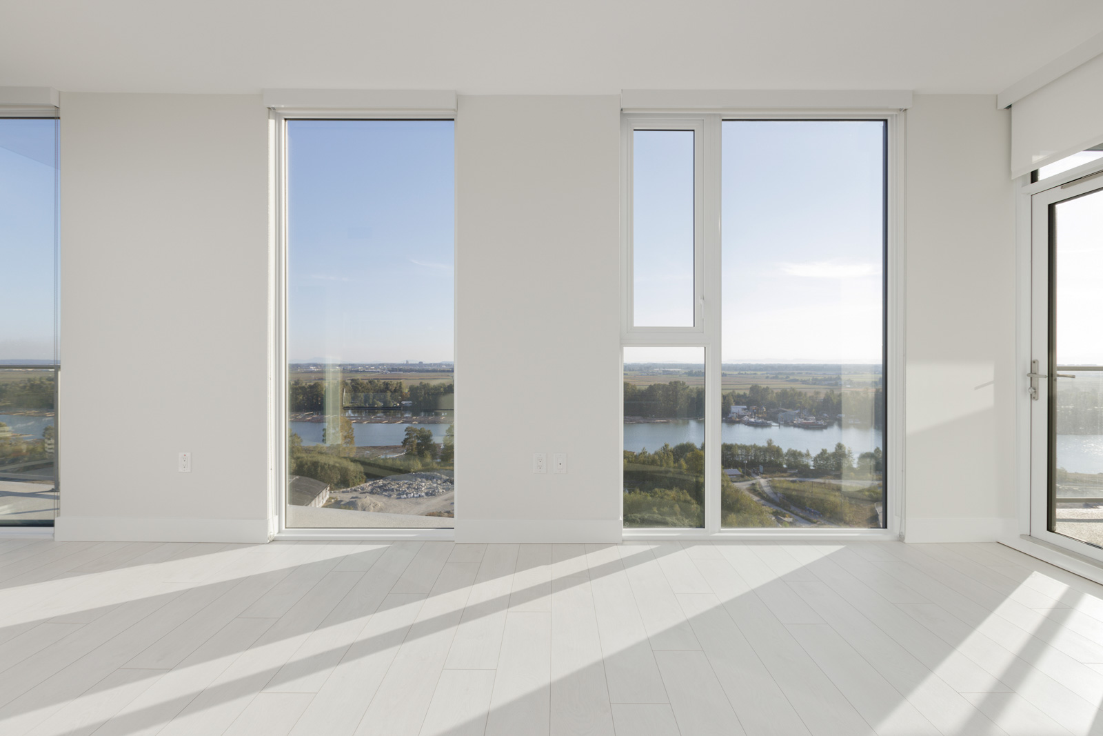 A bright, empty room with large windows offers a view of a river and lush greenery outside. The room has light-colored wooden flooring.