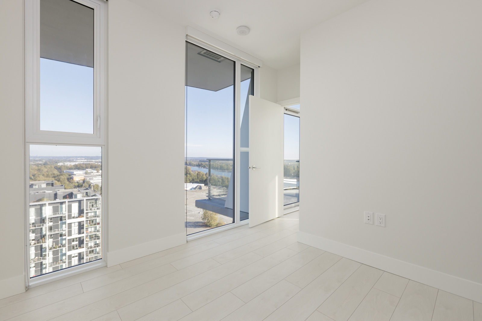 Bright, empty room with light-colored hardwood floors, large windows, and a slightly open door leading to a balcony with a view of buildings and a distant landscape.