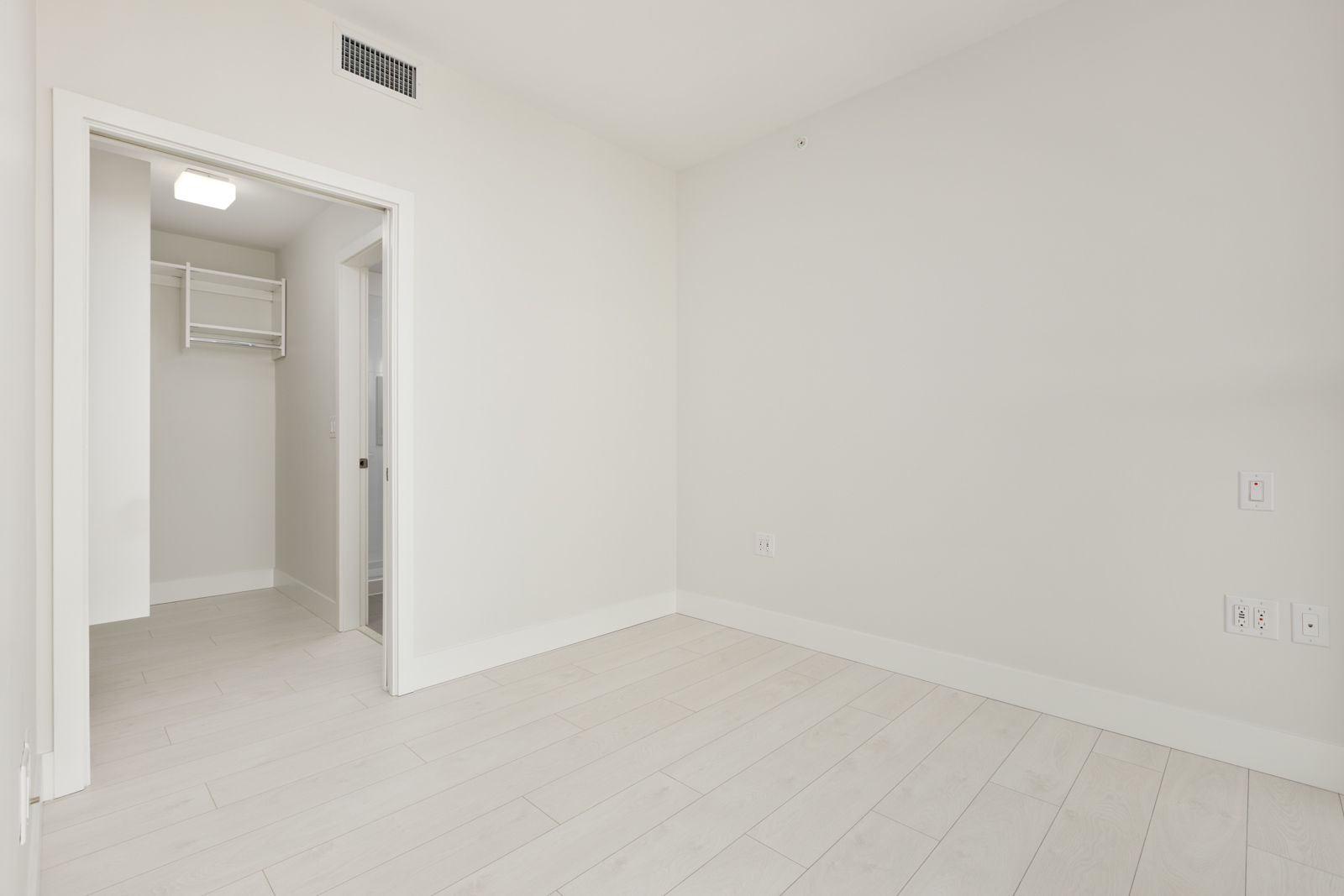 A minimalistic empty room with white walls and light wooden flooring. A walk-in closet with shelving is visible through an open door.