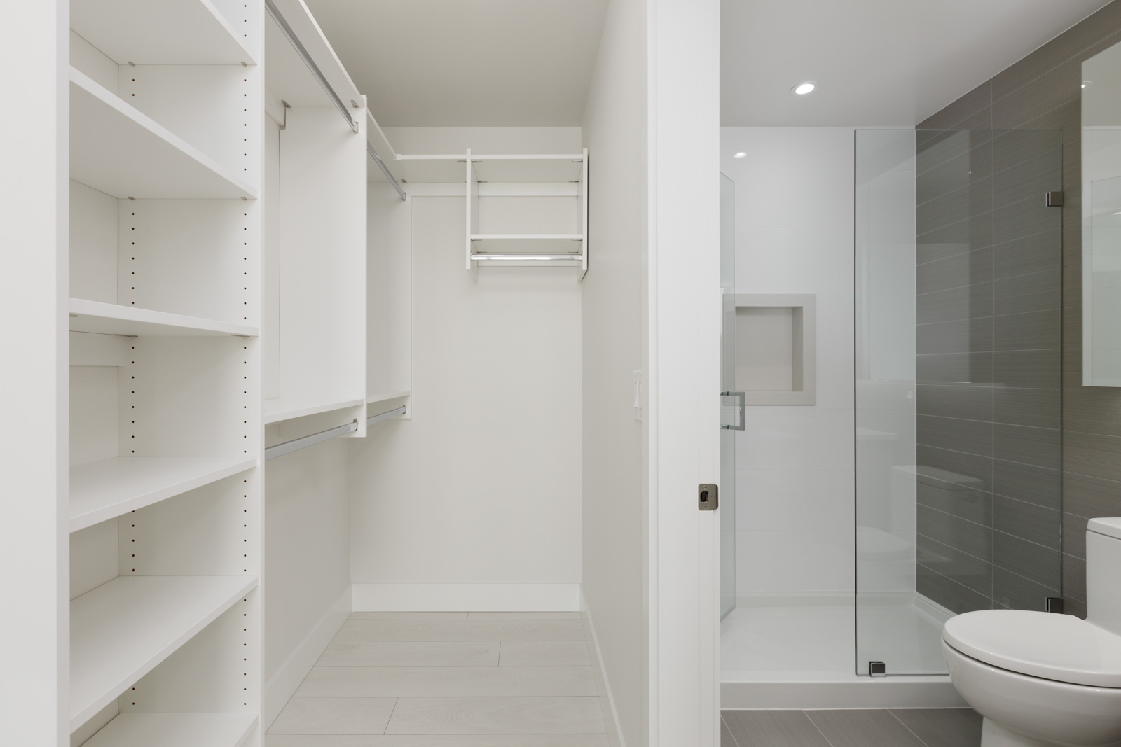 A modern closet with white shelves and hanging rods to the left, adjacent to a bathroom featuring a glass-enclosed shower and a white toilet.
