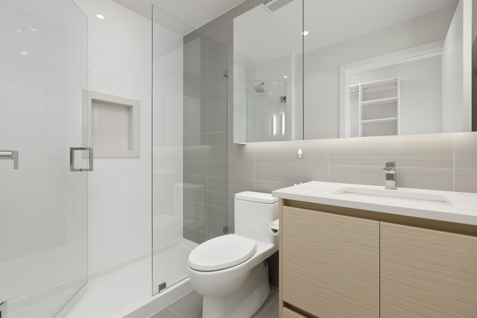 A modern bathroom with glass shower enclosure, built-in shower shelf, toilet, sink, light wood vanity, and large wall mirror. The color palette includes white, beige, and light gray.