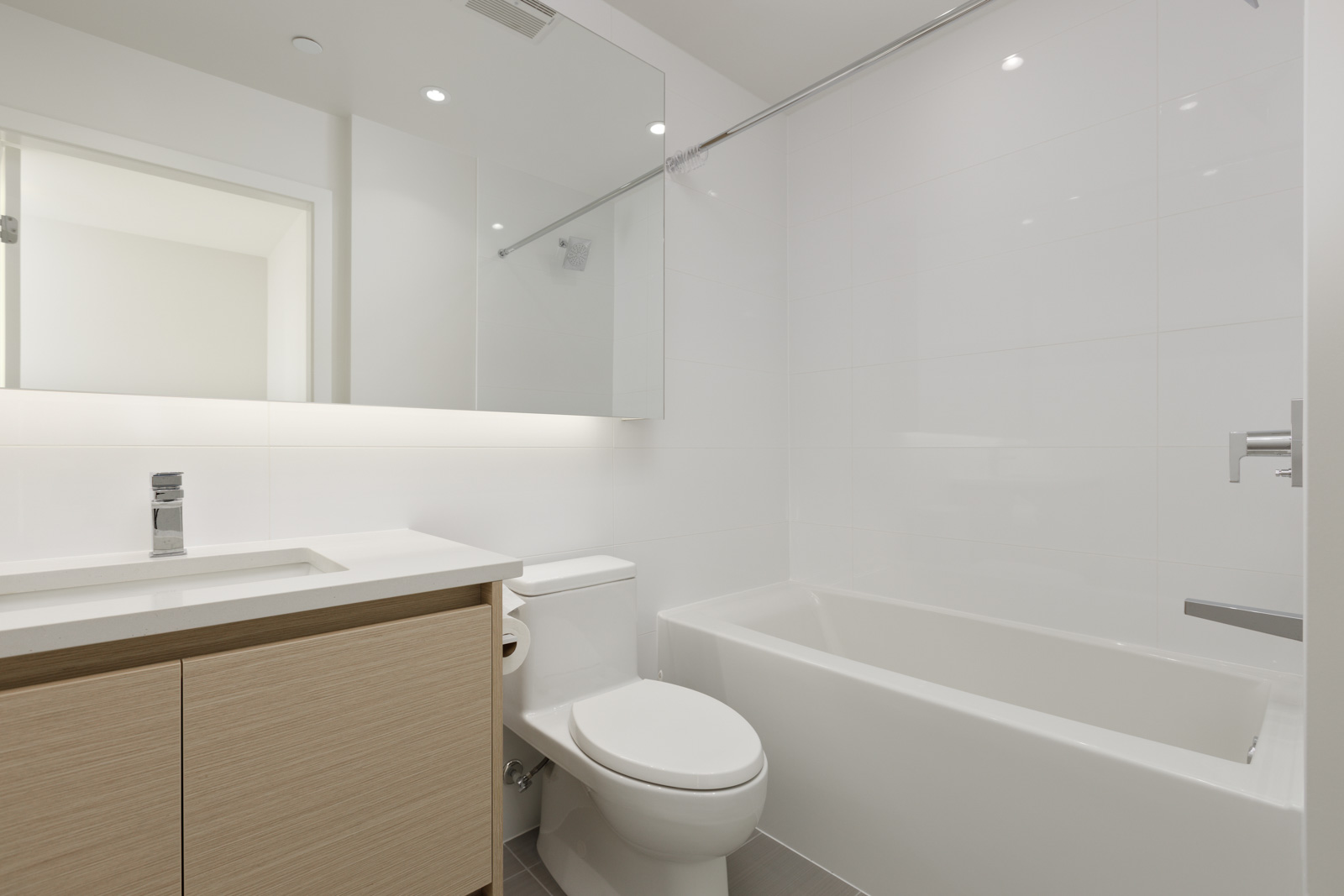 A modern bathroom with a vanity sink, toilet, bathtub, and large mirror. The decor is minimalistic with white walls and light wooden cabinetry.