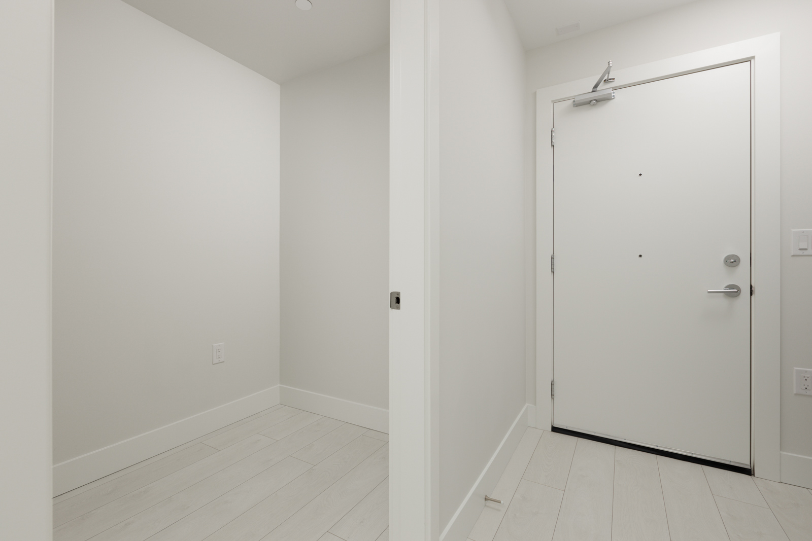 A minimalist white hallway with light wood flooring. The hallway leads to a closed white door with a doorknob and latch on the right.