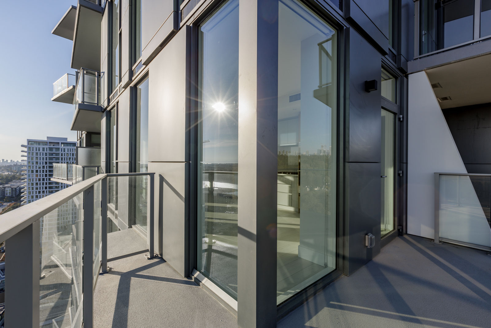 A corner balcony with glass railings and floor-to-ceiling windows in a high-rise building, overlooking a cityscape in the distance.