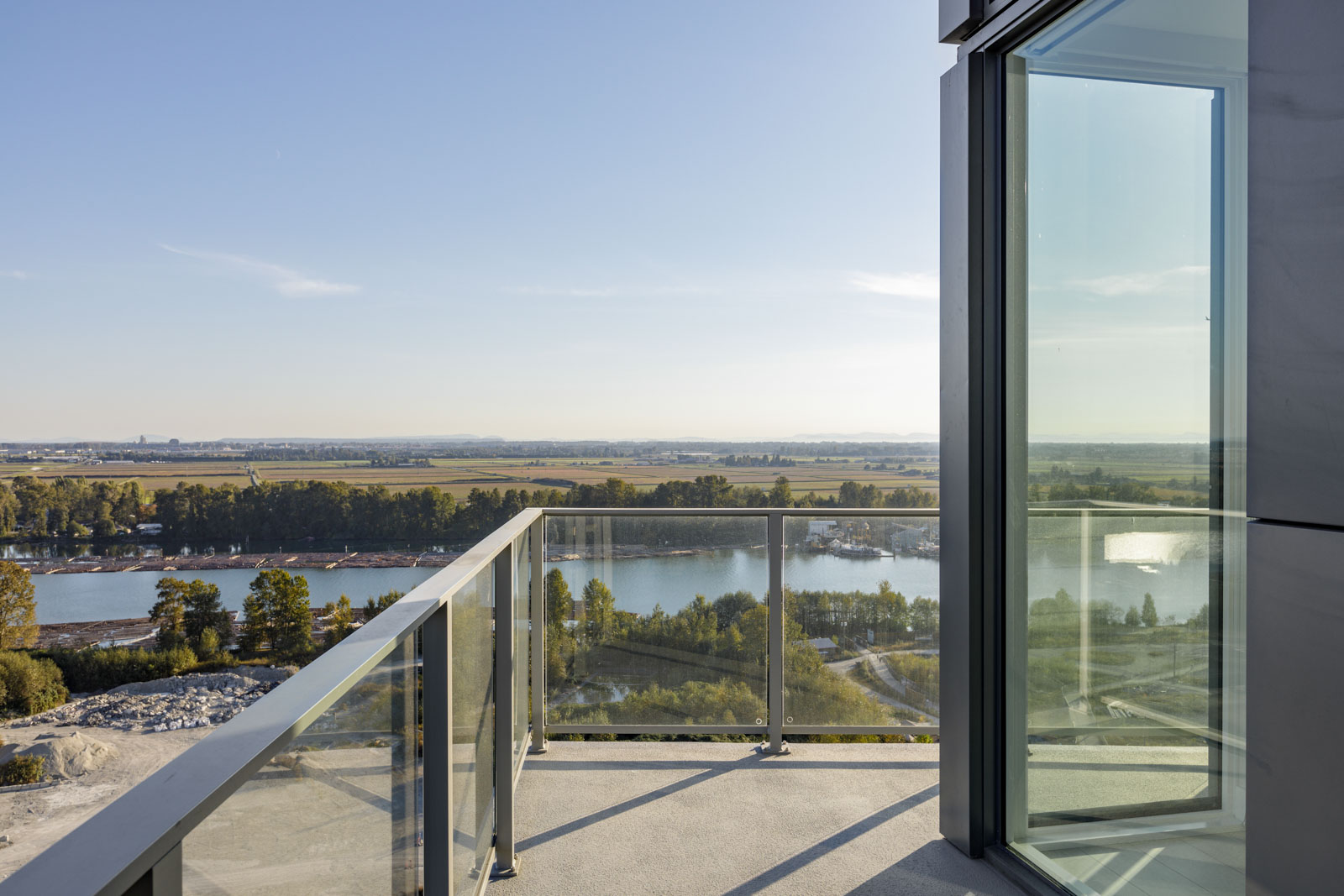 A view from a modern balcony with glass railings, overlooking a river and surrounding greenery under a clear sky.