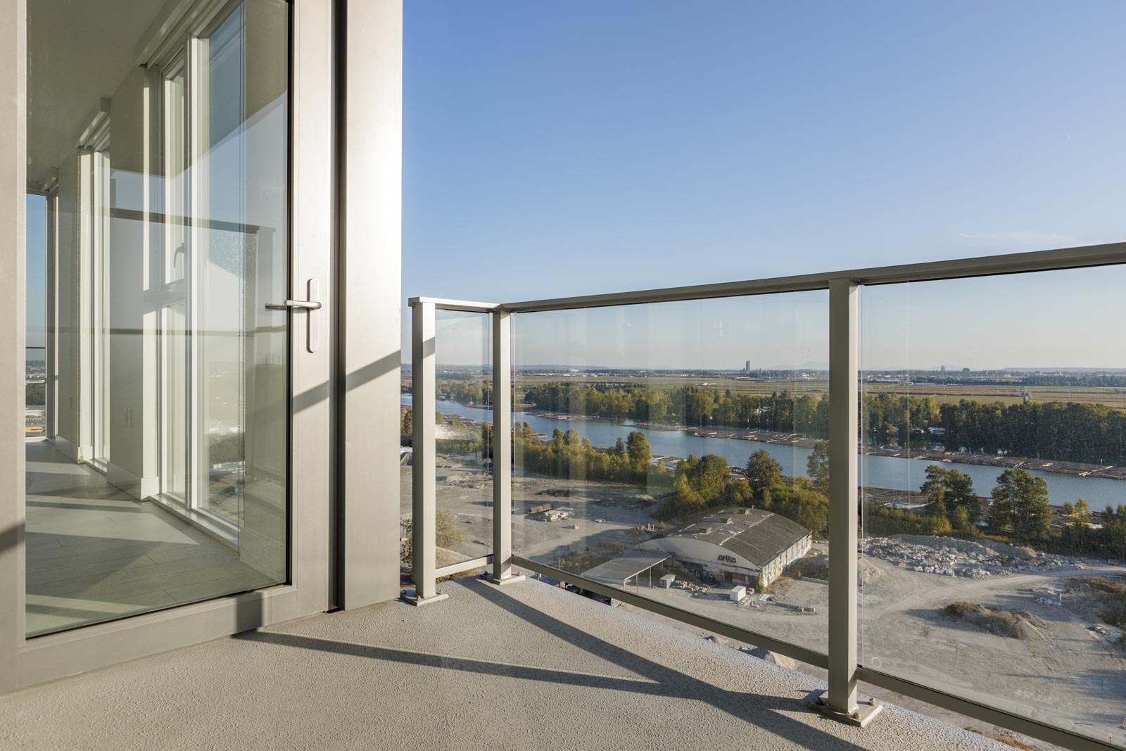 Balcony of a modern building overlooking a river and construction site with glass railing and an open door leading to an indoor area.