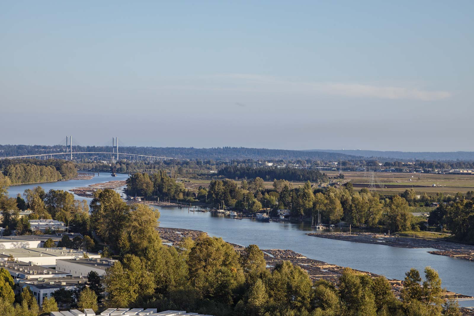 A river winds through a landscape featuring dense trees, a bridge in the distance, and industrial areas along the banks under a clear blue sky.