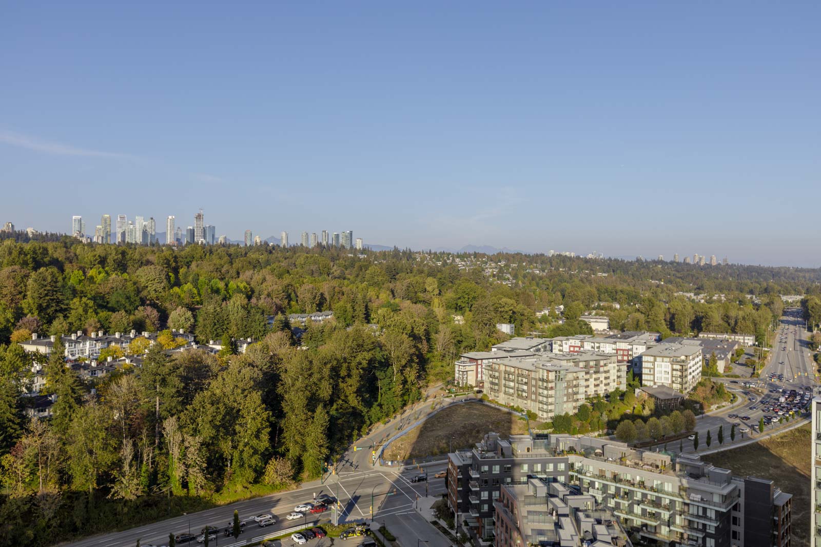 Aerial view of a cityscape featuring high-rise buildings in the distance, a large expanse of trees, residential buildings, and a major roadway with vehicles on the right. Sky is clear and blue.