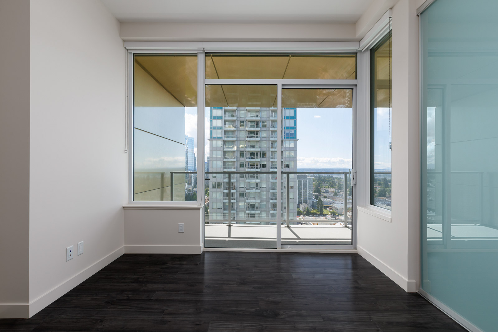 Unfurnished apartment room with dark wood flooring, large glass doors, and windows opening to a balcony with a view of a cityscape and distant mountains.