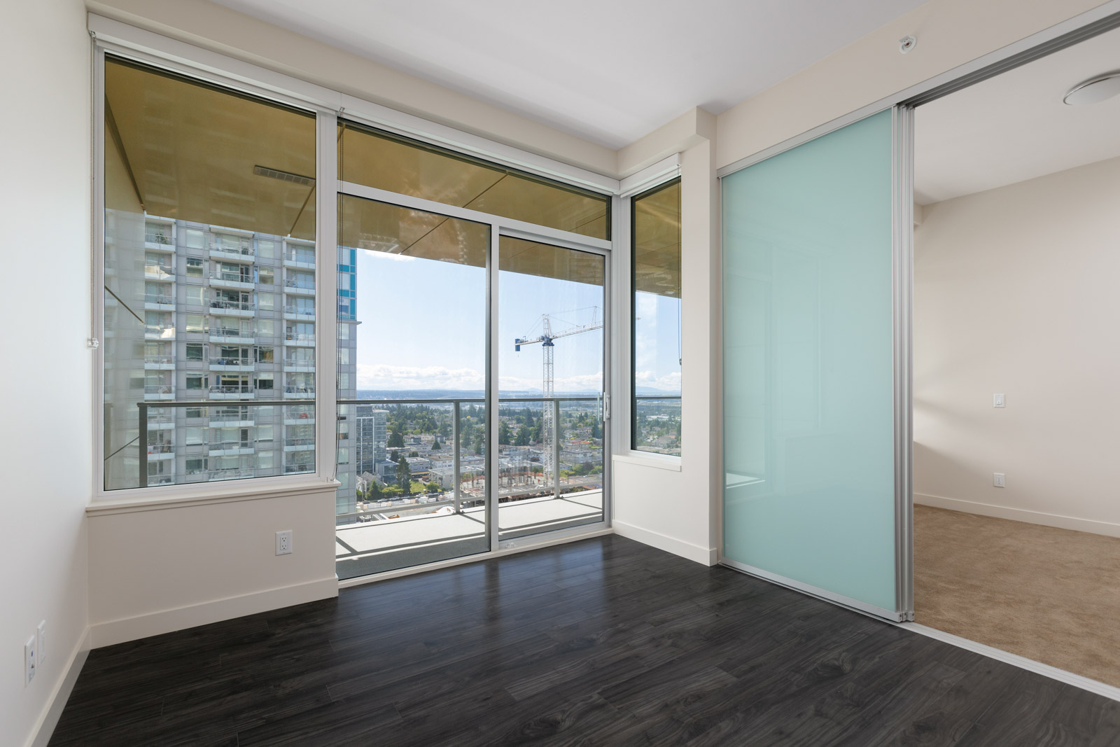 Empty room with dark wood floor, large windows, and a view of buildings and a crane outside. There's a frosted glass sliding door leading to another room with beige carpet.