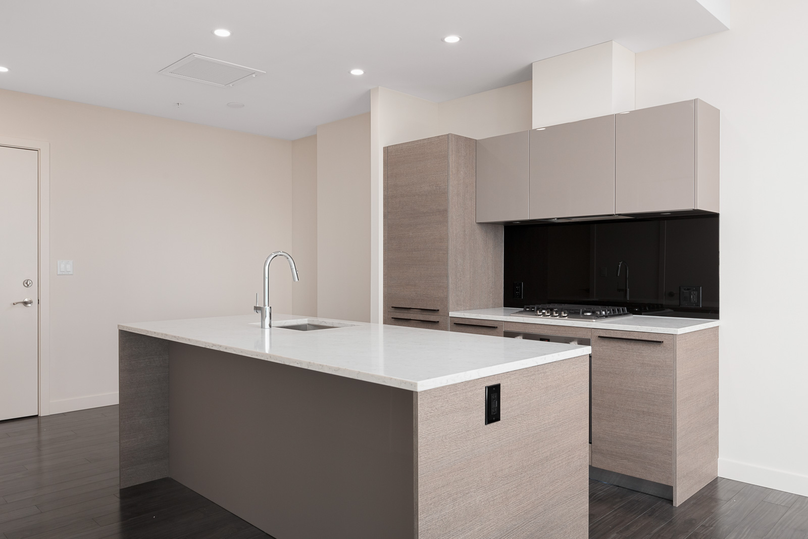 Modern kitchen with a large island, sink, faucet, and beige cabinetry. Black backsplash with a stovetop and vent. Light walls and dark wood flooring.