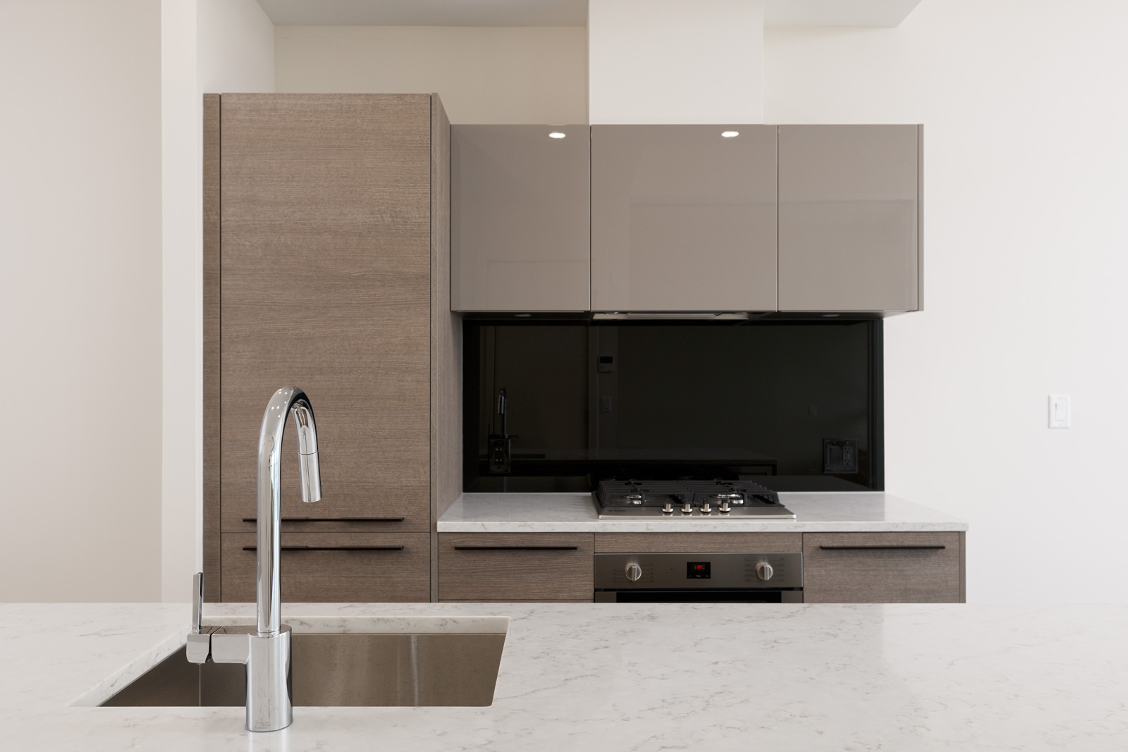 Modern kitchen with light brown cabinets, black glass backsplash, and a built-in gas stove. A chrome faucet is installed on a white countertop with an integrated sink.
